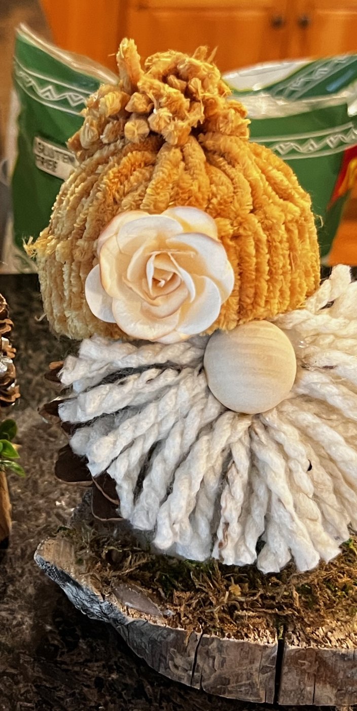 Handmade gnome with a golden chenille hat, a cream rose, and a white yarn beard on a wood slice