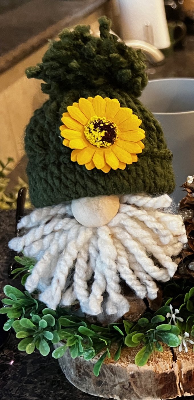 Handmade gnome with a green knit hat, a yellow sunflower, and a white yarn beard on a wood slice