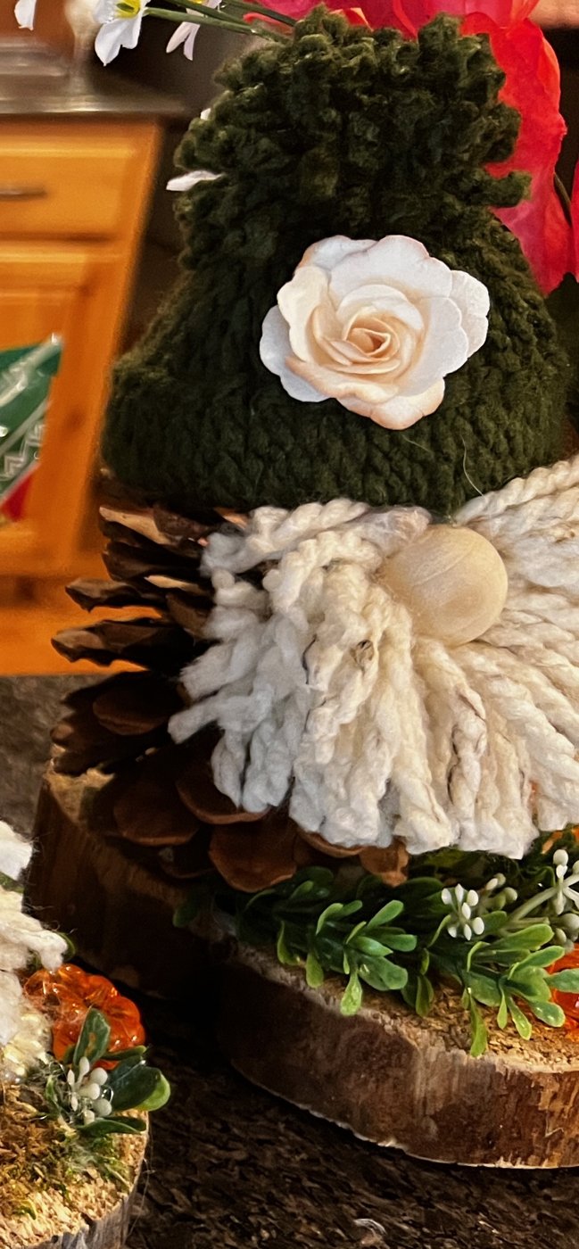 Handmade gnome with a green hat, a white rose, and a layered pinecone body on a wood slice